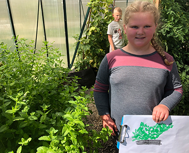 Female student holds up a piece of paper with artwork on it in a greenhouse